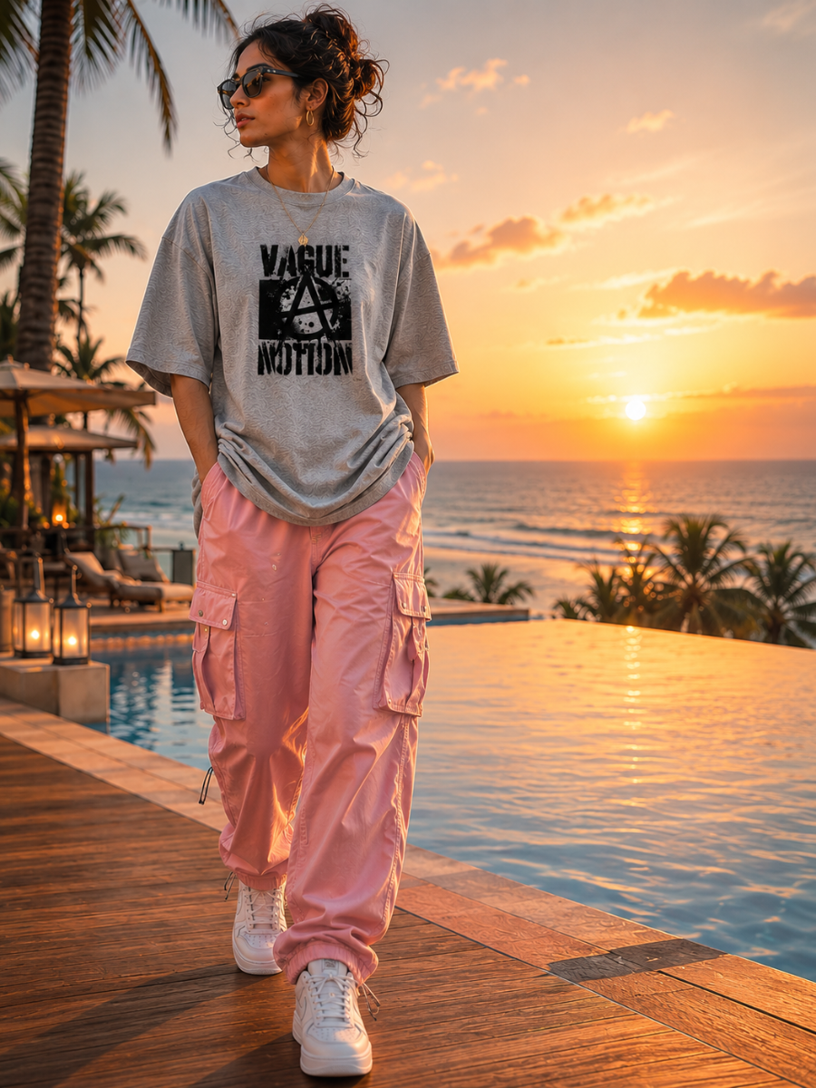 female model wearing a gray zingleberyy's oversized unisex t-shirt with text and pink cargo pants standing by a pool with a sunset view.