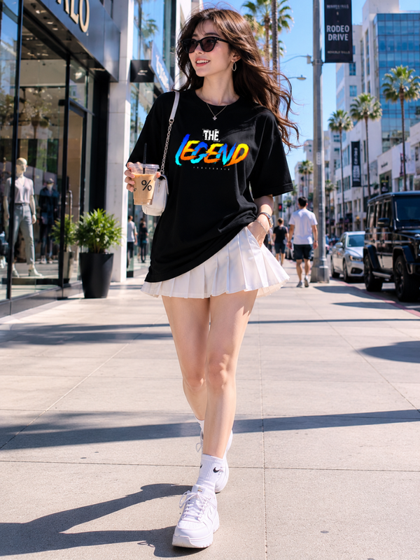 Female model wearing Zingleberry Premium Unisex Classic Crew The Legend black graphic T-shirt paired with a white pleated skirt in a modern city street fashion photoshoot.