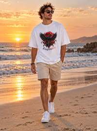 Man wearing Zingleberry Blood Moon Raven white oversized cotton T-shirt with beige shorts at sunset beach.