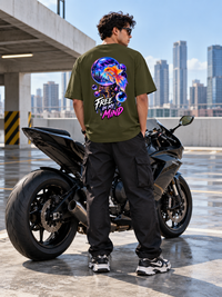 Male model wearing Zingleberry Free In My Mind olive green oversized T-shirt with vibrant cosmic goldfish back artwork, black cargo trousers and sneakers beside a sports motorcycle