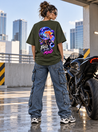 Female model wearing Zingleberry Free In My Mind olive green oversized T-shirt with vibrant goldfish cosmic back artwork, blue cargo jeans and sneakers in urban rooftop setting