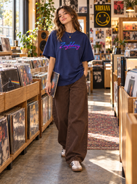 Female model wearing Zingleberry royal blue oversized T-shirt with neon signature logo, brown wide-leg jeans and casual sneakers inside a vintage record store.