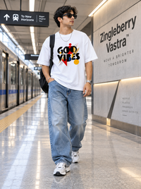 Male model wearing Zingleberry Good Vibes white oversized abstract graphic T-shirt with blue baggy jeans and sneakers at a modern metro station.