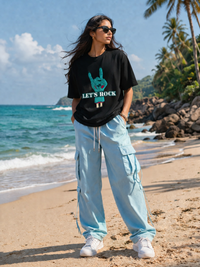 Female model wearing Zingleberry Let's Rock Premium Black Cotton Graphic T-Shirt with light blue cargo pants at a beachside location.