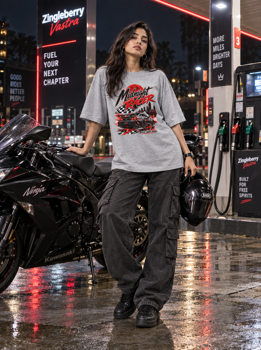 Female model wearing Zingleberry Midnight Racer Grey Melange oversized racing car graphic T-shirt with black cargo pants beside a sport motorcycle at a night fuel station.