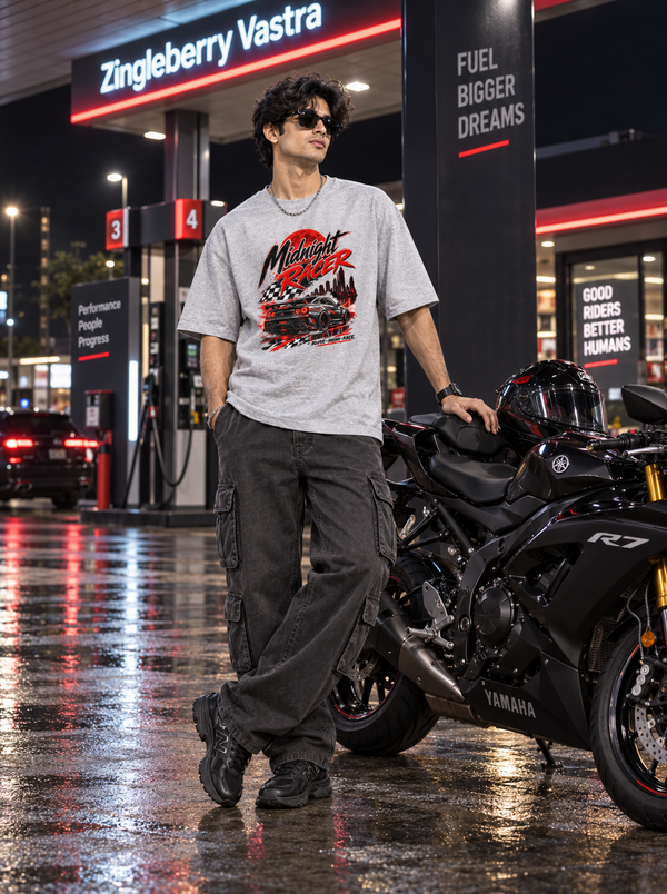 Male model wearing Zingleberry Midnight Racer Grey Melange oversized automotive graphic T-shirt with dark cargo pants beside a sport motorcycle at a night fuel station.