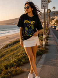 Female model wearing Zingleberry Premium Unisex Classic Crew Born Rage black graphic T-shirt styled with a white skirt in a beachside fashion photoshoot.