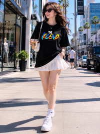 Female model wearing Zingleberry Premium Unisex Classic Crew The Legend black graphic T-shirt paired with a white pleated skirt in a modern city street fashion photoshoot.