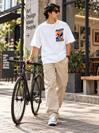 Male model wearing Zingleberry Progress Over Perfection white oversized graphic T-shirt with small front chest artwork, beige cargo trousers and white sneakers beside a bicycle.