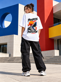 Female model wearing Zingleberry Progress Over Perfection white oversized T-shirt featuring bold geometric staircase back artwork with black cargo trousers at a modern architectural location.