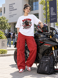 Female model wearing Zingleberry Racing Legend white oversized racing graphic T-shirt with red cargo pants beside a sport motorcycle on a college campus.